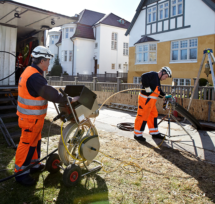 sewer ninjas crew members performing a drain cleaning service in the backyard of someone's home. men are wearing orange jumpsuits and hard helmets with a large black pipe coming out of a sewer hole